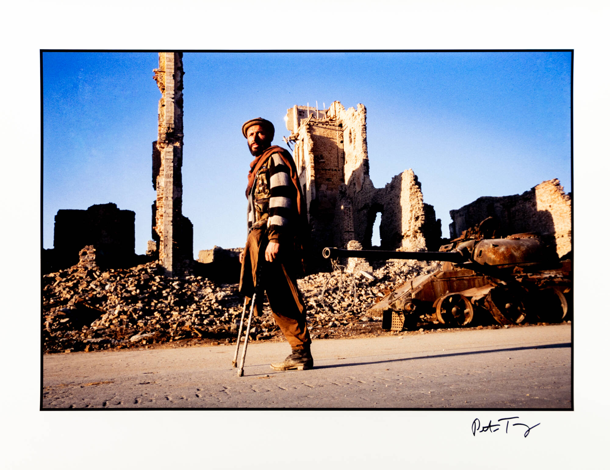 A horizontal color photograph of a man standing on crutches. Behind the man are the ruins of some buildings and a rusty tank.